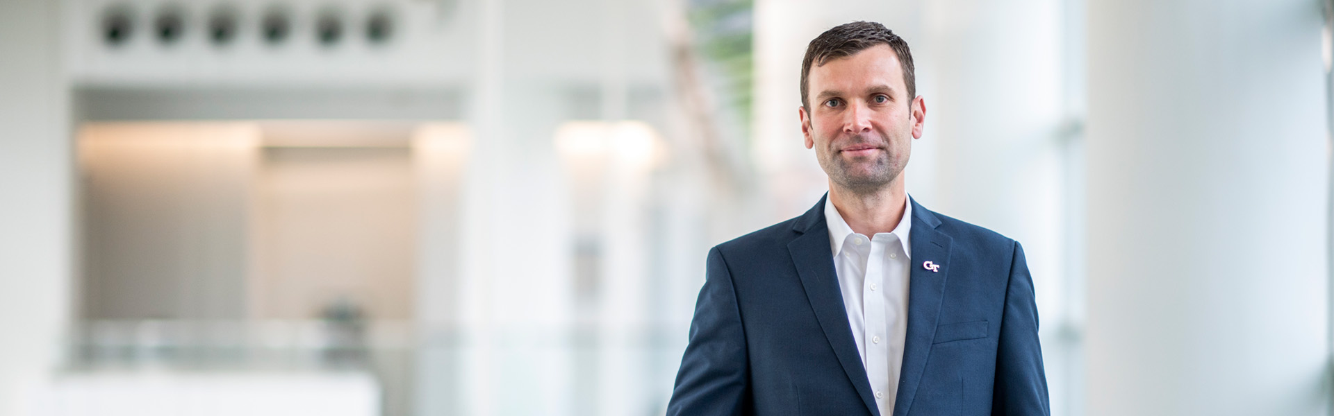 Man in a blue suit jacket smiling while leaning on a glass railing in a modern building
