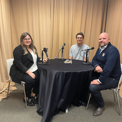 Three individual sitting a table with microphones, recording a podcast