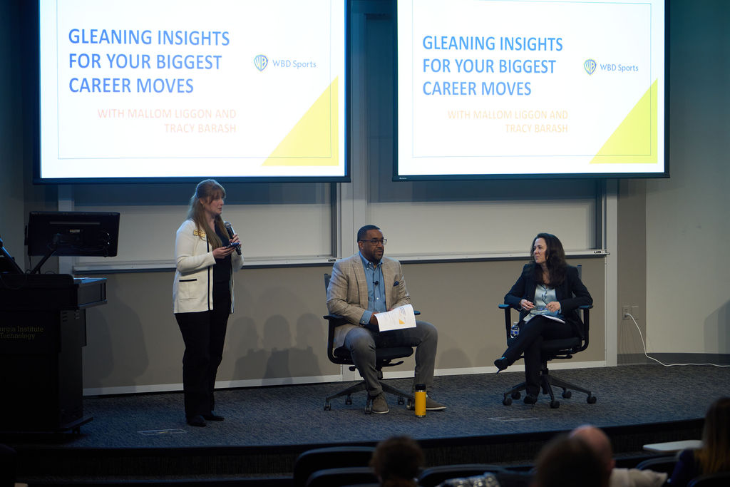 One female presenter is standing on a stage next to two seated panelists. Behind them is a presentation that reads “Gleaning Insights For Your Biggest Career Moves.”