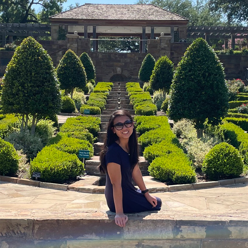 Sky Blevins, Evening MBA ’23, smiles sitting on a stone wall in a garden  