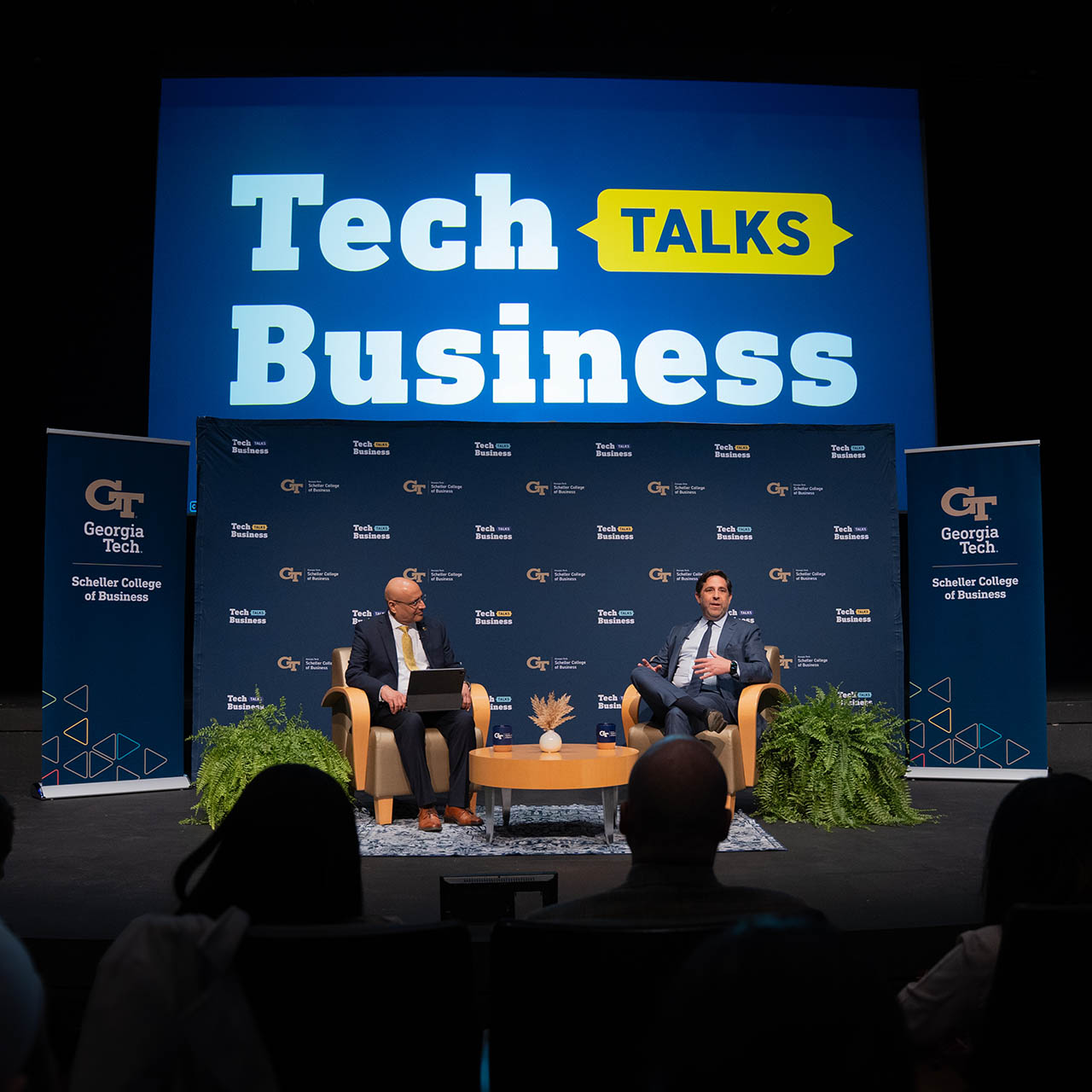 Two men in suits sit in chairs on a stage