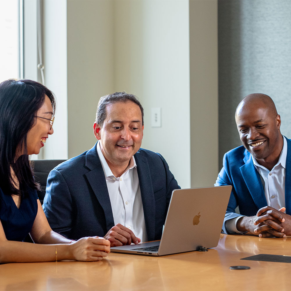 Three people sit at a conference table working on laptops, engaged in discussion in a modern office setting.