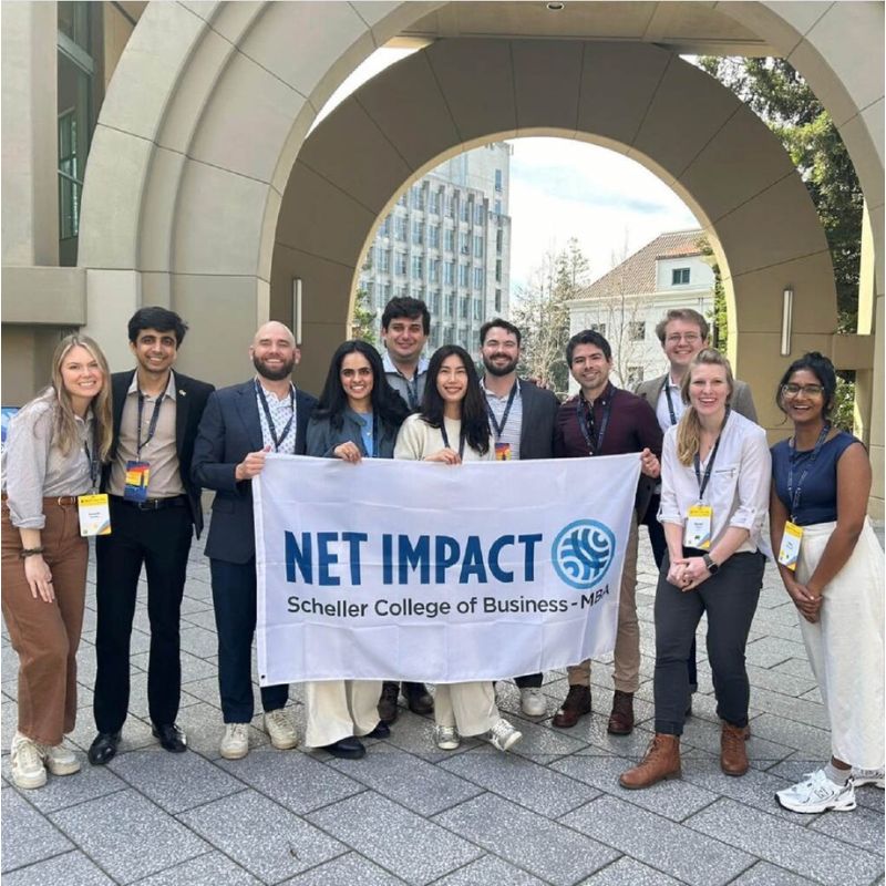 Scheller MBA students and staff standing outside holding a white banner with "Net Impact" written on it